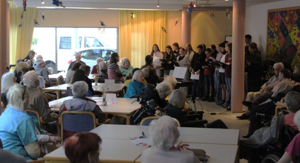 BLOG-Beitrag
FES-Schüler überraschen Bewohner im Altenzentrum musikalisch
Tränen der Rührung, Glück und Freude: So viele gute Gefühle in einem Gesicht ablesen zu können, ist wohl der beste Beweis für eine gelungene Aktion.
20 Schülerinnen und Schüler unseres FES-Jugendchors blickten bei ihrem kleinen musikalischen Ständchen im Eugen-Lederer-Altenzentrum in HDH am Mittwoch in ganz viele glückliche Gesichter: Die Schüler unseres FES-Gymnasiums und unserer FES-Realschule besuchten mit FES-Chorleiter Daniel Kindsvater und Schulleiter Andreas Borkeloh die Senioren im AWO-Altenzentrum in der Stadtmitte und überraschten mit europäischer Chormusik unterschiedlicher Stilistik aus dem aktuellen Konzertprogramm. Die Cafeteria war beim Besuch der jungen Sänger aus den Klassenstufen 7-10 proppenvoll gefüllt und die jungen Choristen erfüllten das Auditorium mit fröhlicher Musik. Besondere Freude bereitete den älteren Menschen, dass zum Abschluss des musikalischen Ständchens gemeinsam gesungen wurde. „Vielen Dank, dass Ihr heute bei uns seid. Unsere Bewohner freuen sich riesig, dass ihr für sie singt“, bedankte sich Nadine Schnell, Pflegedienstleitung im AWO-Altenzentrum am Ende des gut 30-minütigen kleinen chorischen Vortragsprogramms.